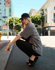 A model crouching on the street wearing a gray 'Kindness Club' t-shirt, black leggings, black slides, and a black dad cap with white 'I I II I' embroidery