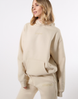 Side-angled-view of a model wearing a beige hooded sweatsuit with 'LEGEND' in tonal printing on the center of the chest on top left thigh on a white background