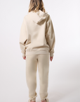 Back-view of a model wearing a beige hooded sweatsuit in front of a white background