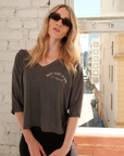 Model wearing a one-size, dark gray v-neck top with 'Not Just A Girl A Legend' printed in white on the left side of the chest paired with black leggings and sunglasses in front of a window with a city view.