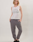 Model wearing a tank top with "Make Your Own History' printed in gray paired with gray sweatpants and black open-toed slides on a plain background