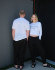 Two models standing against a plain wall, the model on the left posed giving a back-view featuring a a white, tonal print, short sleeve t-shirt with graphic art that says "Space." across the back of the t-shirt paired with black pants, the model on the right is wearing a white cropped, long-sleeve t-shirt featuring graphic art that says 'In My Fearless Era' in gray puff-print with black sweat pants