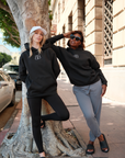 Two models wearing BB logo oversized sweatshirts, the model on the left paired the sweatshirt with black leggings and a white dad cap featuring black 'I I II I' embroidery and the model on the right paired the sweatshirt with sunglasses and gray joggers casually posing on the street in Los Angeles