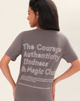 Close-up, back-view of a model wearing a one-size gray, short-sleeve t-shirt that has white graphic art that says 'The Courage, Authenticity and Magic club' on the back with black leggings in front of a white background