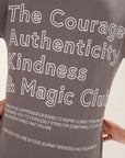 Model wearing a gray t-shirt with motivational text printed in white that says "The Courage Authenticity And Magic Club" on the back in front of a white background