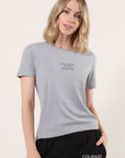 Model wearing a gray crew neck t-shirt with the mantra 'COURAGE OVER COMFORT' printed in black on the center of the chest paired with 'Courage Over Comfort' black pants with white print in front of a white background