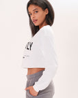 Side-view of a model wearing a long sleeve white boxy tee that says "Imperfectly Perfect" in black graphic art in front of a white backdrop.