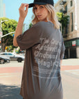 Close-up, back-view of a model wearing a one-size gray, short-sleeve t-shirt that has white graphic art that says 'The Courage, Authenticity and Magic club' on the back with black leggings and a black dad cap with white I I II I embroidery posing on the street in Los Angeles
