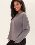 Model is wearing an oversized long sleeve gray t-shirt with a crew neck and white printed BB logo in the center of the chest and Birdiebee printed on the wrist of one sleeve with black leggings, leaning at a side-angle in front of a white backdrop