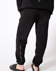 Back-view of a model wearing black sweatpants with white 'Own Your Moment' text on a white background