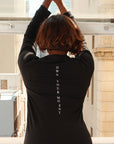 Model posing featuring a back-view of a long sleeve barely-sheer black burnout t-shirt with a 'Own Your Moment' printed in white down the center of the back, paired with black active shorts leaning on a brick wall next to windowsring