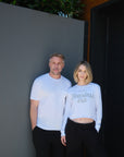 Two models standing against a dark wall, the model on the left is wearing a white, tonal, short-sleeve t-shirt that says 'Personal.' on the front left, chest pocket and the model on the right is wearing a white, long-sleeve, cropped t-shirts that says 'In My Fearless Era' printed in gray, both paired with black joggers