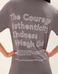 Model wearing a gray t-shirt with motivational text printed in white that says "The Courage Authenticity And Magic Club" on the back in front of a white background