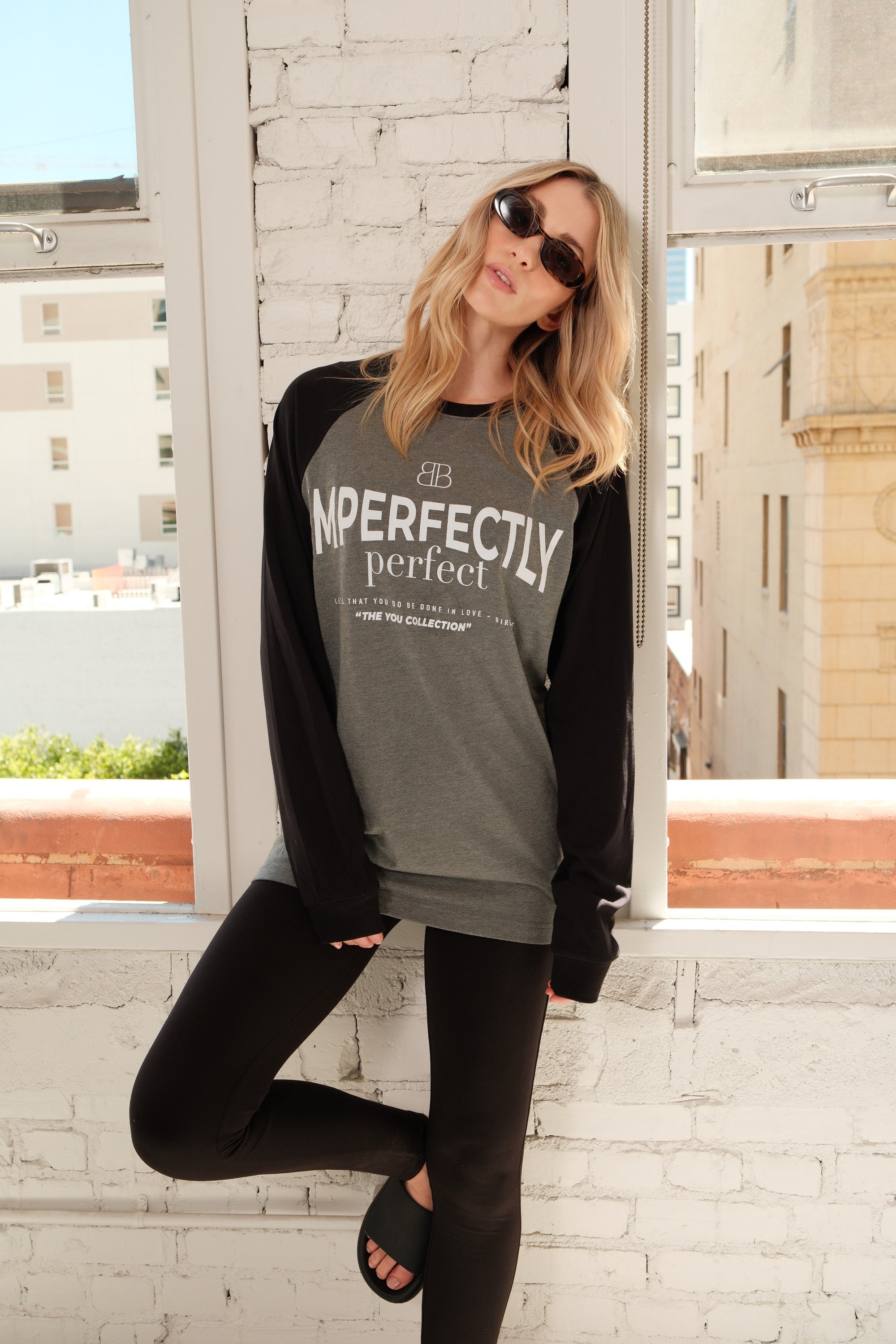 Model wearing a gray shirt with 'IMPERFECTLY Perfect' screenprint standing in front of a window with cityscape background.