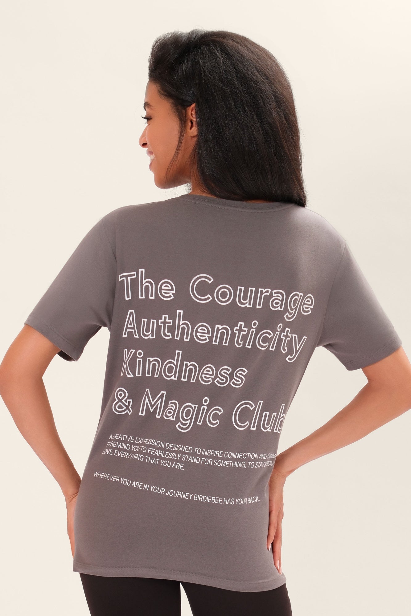 Close-up,  back-view of a model wearing a one-size gray, short-sleeve t-shirt that has white graphic art that says 'The Courage, Authenticity and Magic club' on the back with black leggings in front of a white background