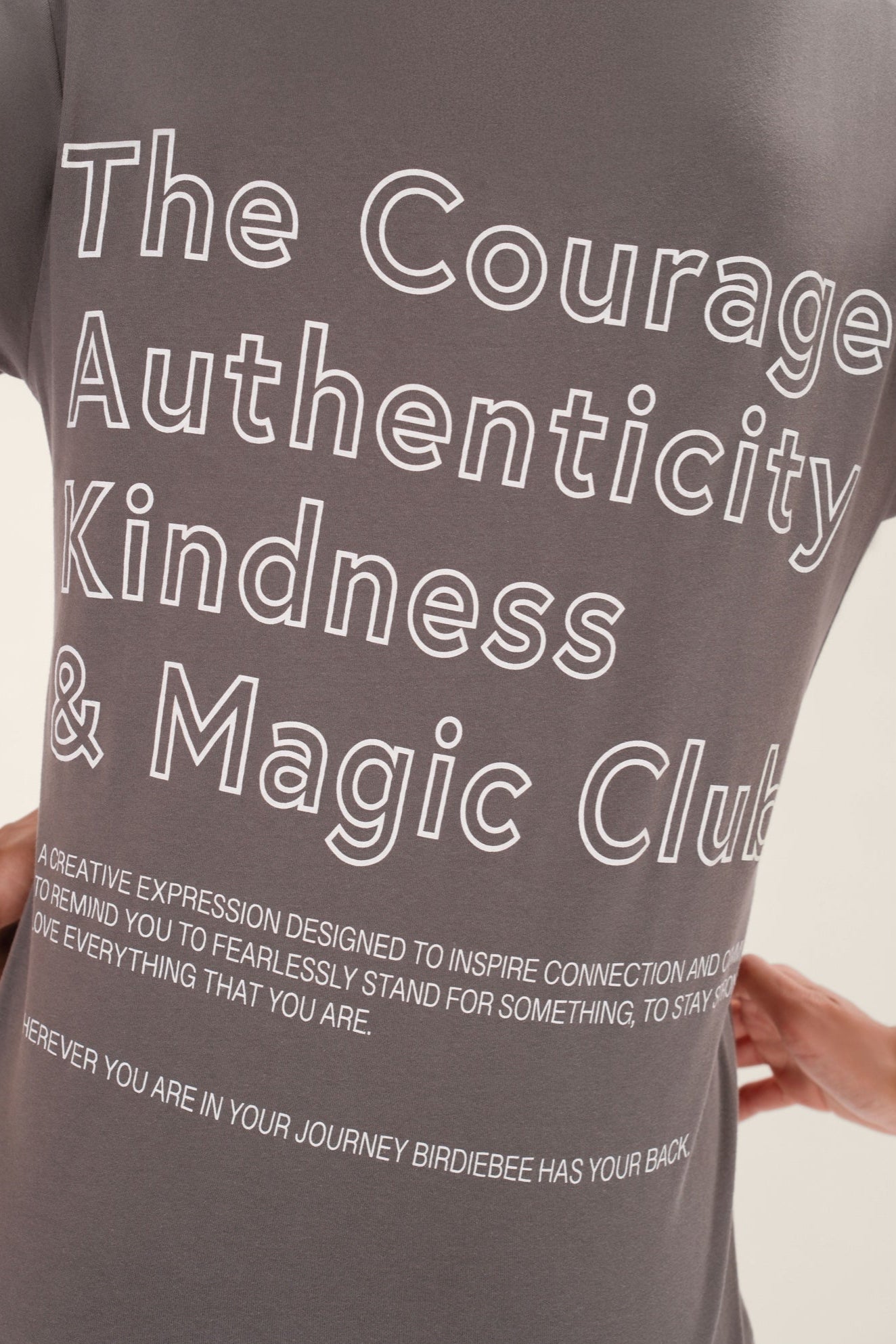 Model wearing a gray t-shirt with motivational text printed in white that says &quot;The Courage Authenticity And Magic Club&quot; on the back in front of a white background