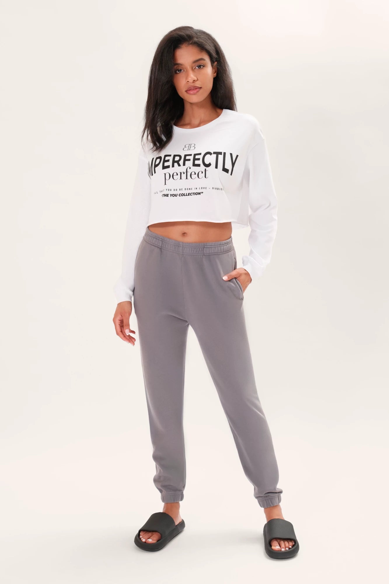 Model wearing a long sleeve white boxy tee that says "Imperfectly Perfect" in black graphic art with gray joggers and black slides in front of a white backdrop.
