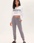Model wearing a long sleeve white boxy tee that says "Imperfectly Perfect" in black graphic art with gray joggers and black slides in front of a white backdrop.