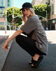 Side-view of a model wearing a gray t-shirt with motivational text printed in white that says "The Courage Authenticity And Magic Club" paired with black leggings and open-toed slides with a black with white embroidery "I I II I" Dad Cap posing bending down on tip-toes on street in Los Angeles