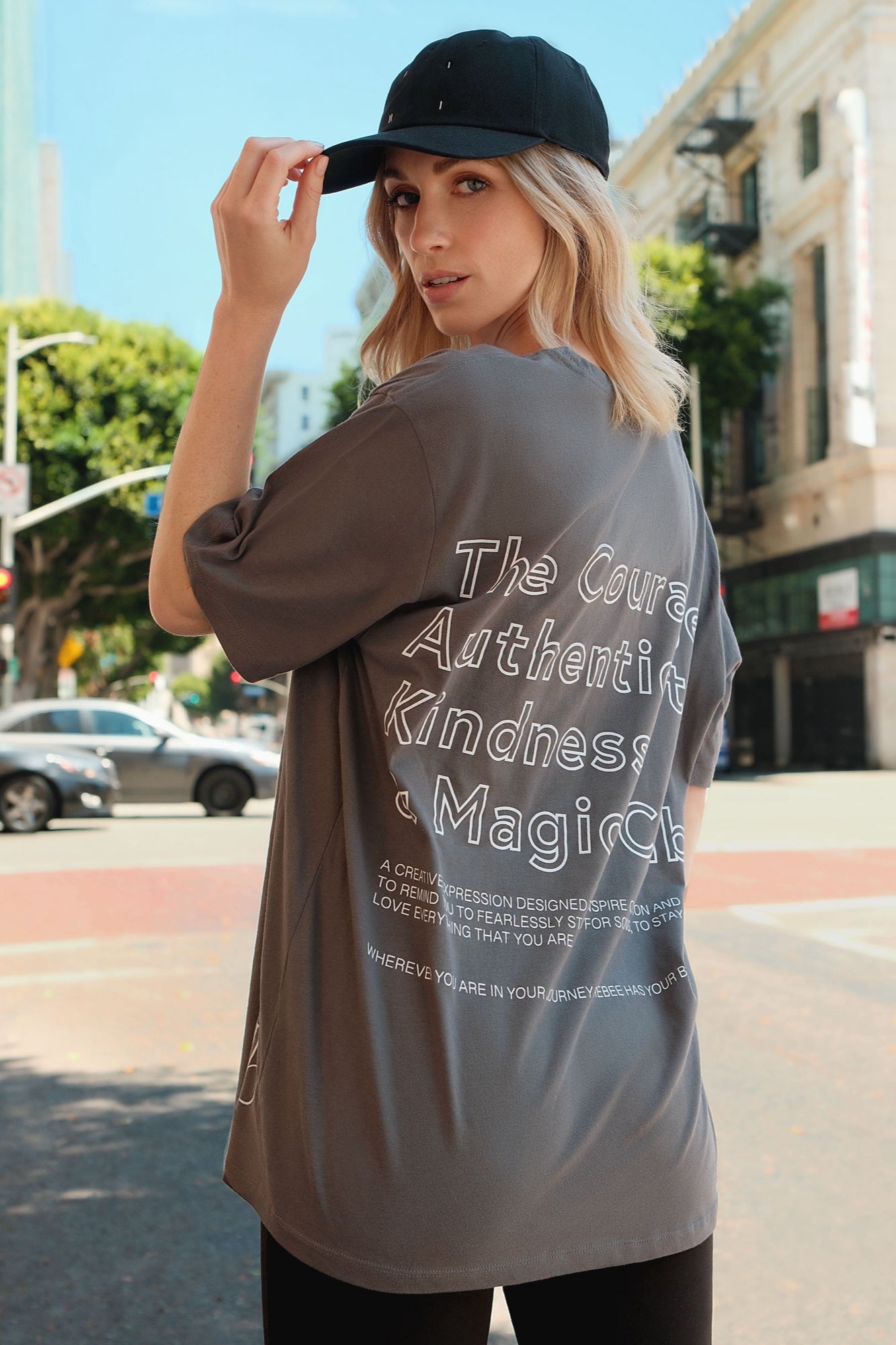 Close-up,  back-view of a model wearing a one-size gray, short-sleeve t-shirt that has white graphic art that says 'The Courage, Authenticity and Magic club' on the back with black leggings and a black dad cap with white I I II I embroidery posing on the street in Los Angeles